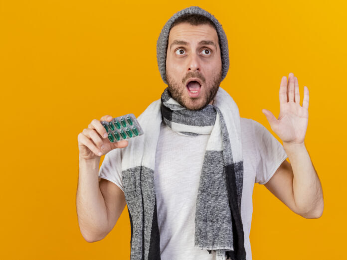 surprised young ill man wearing winter hat and scarf holding pills and showing stop gesture isolated on yellow background