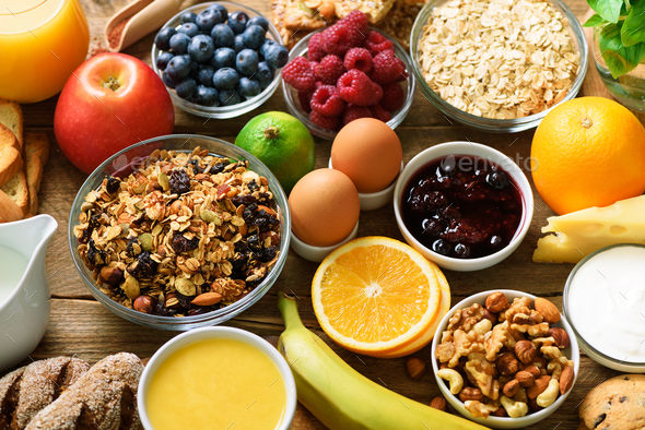 Healthy breakfast ingredients, food frame. Granola, egg, nuts, fruits, berries, toast, milk, yogurt, orange juice, cheese, banana, apple on wooden rustic background, top view, copy space.